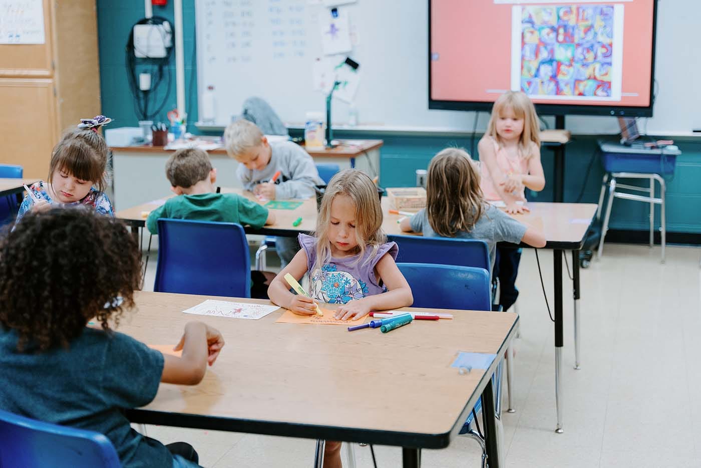 Students in classroom, sitting at tables, reading and coloring.