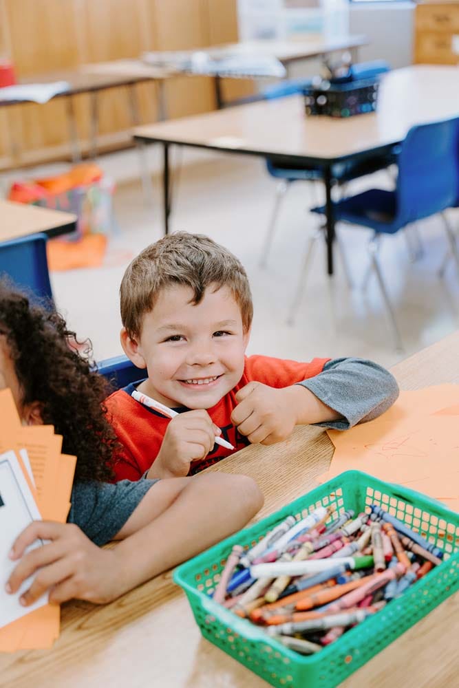 Students in classroom, sitting at tables, reading and coloring.