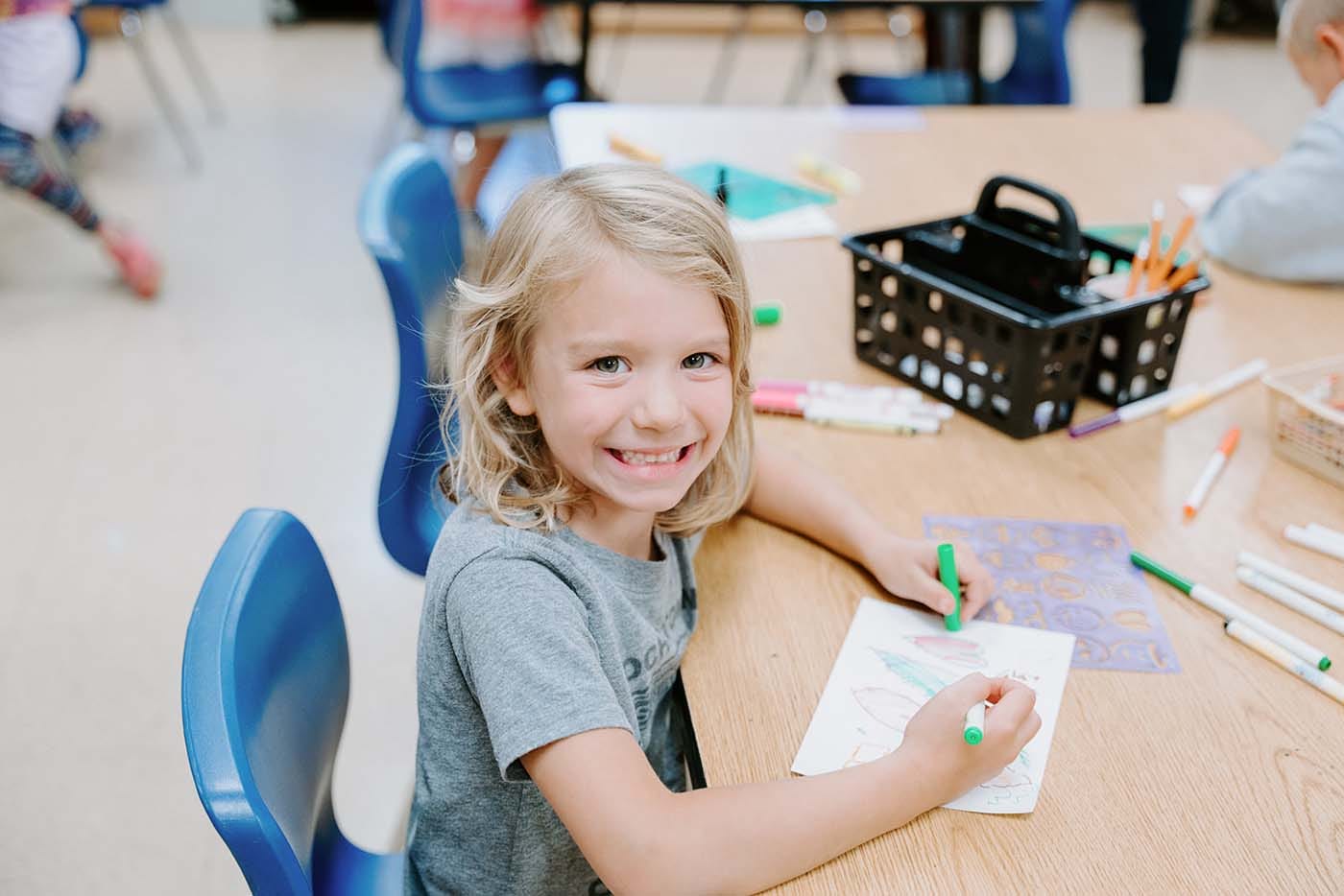 Students in classroom, sitting at tables, reading and coloring.
