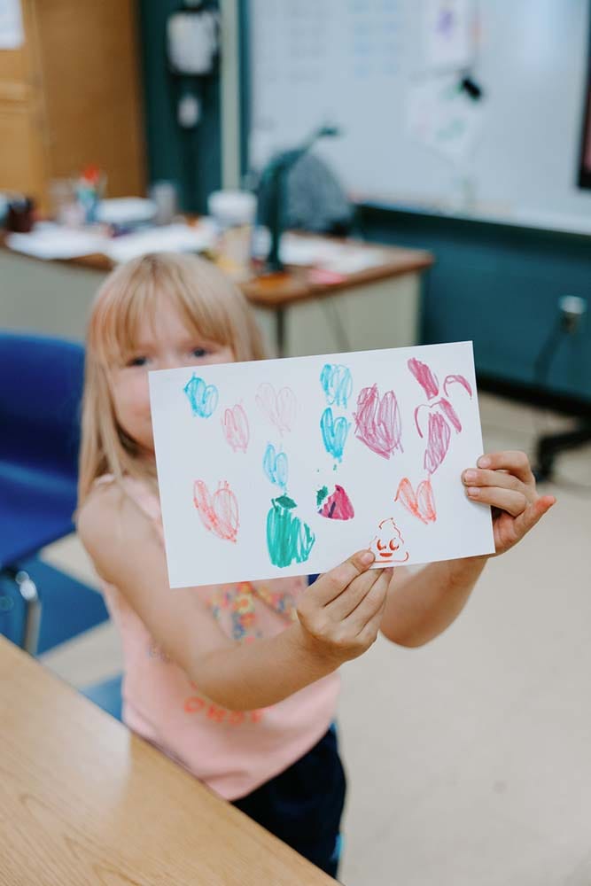 Students in classroom, sitting at tables, reading and coloring.