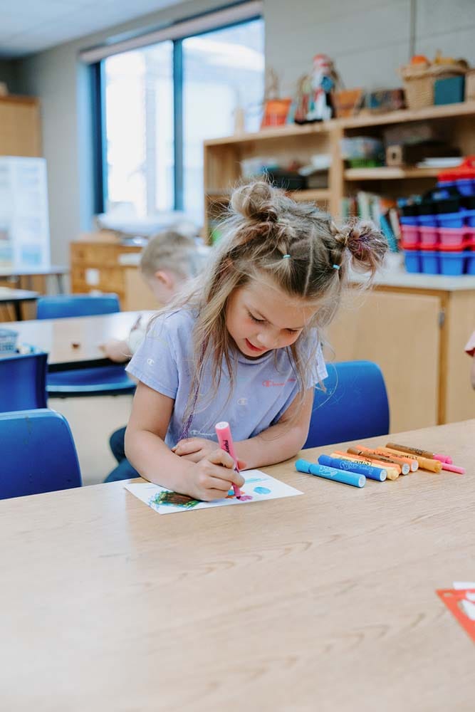 Students in classroom, sitting at tables, reading and coloring.