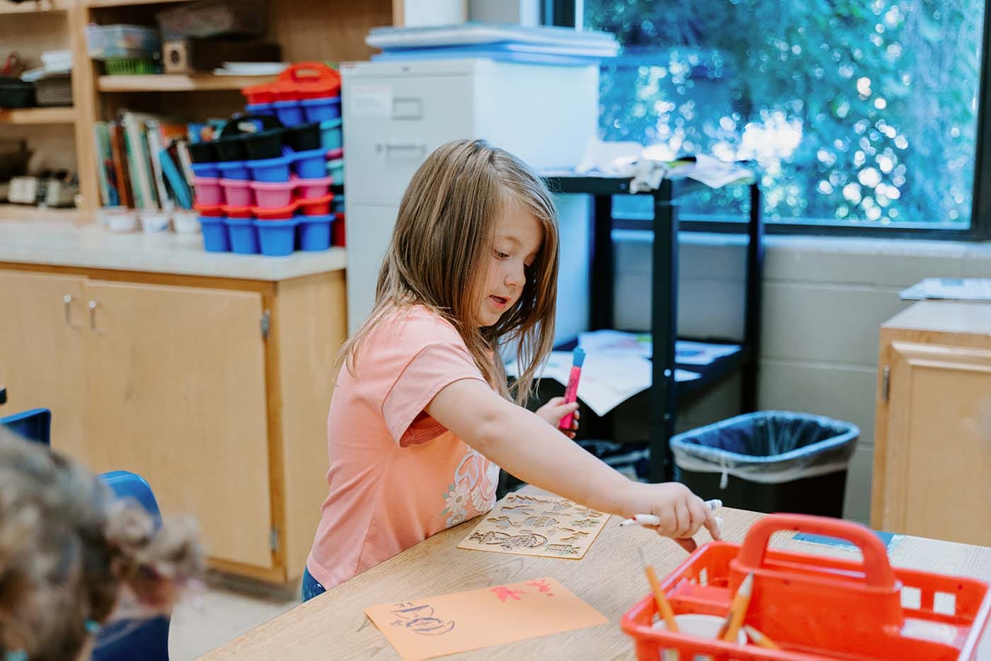 Students in classroom, sitting at tables, reading and coloring.