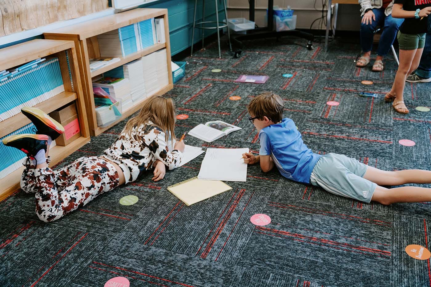 Students in classroom, sitting at tables, reading and coloring.