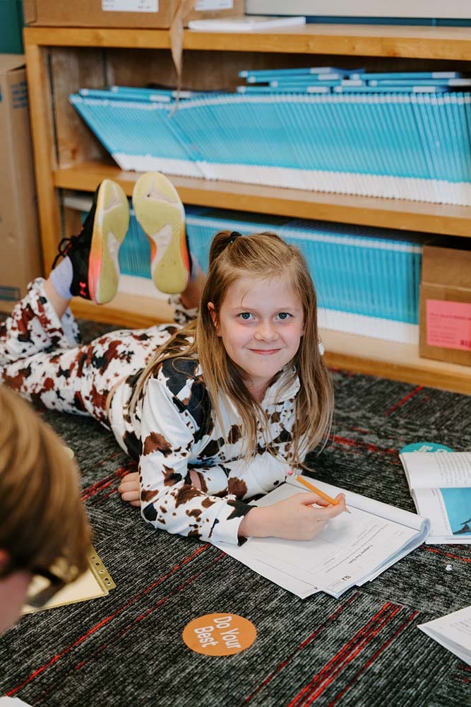 Students in classroom, sitting at tables, reading and coloring.