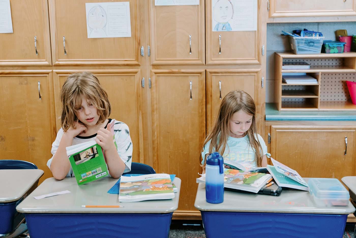Students in classroom, sitting at tables, reading and coloring.