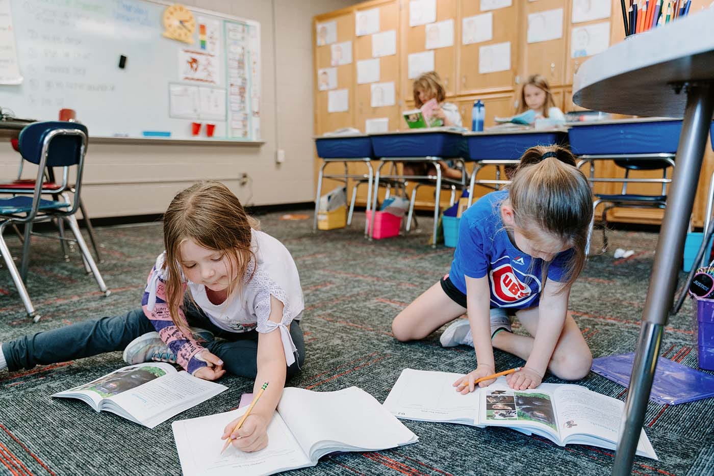 Students in classroom, sitting at tables, reading and coloring.