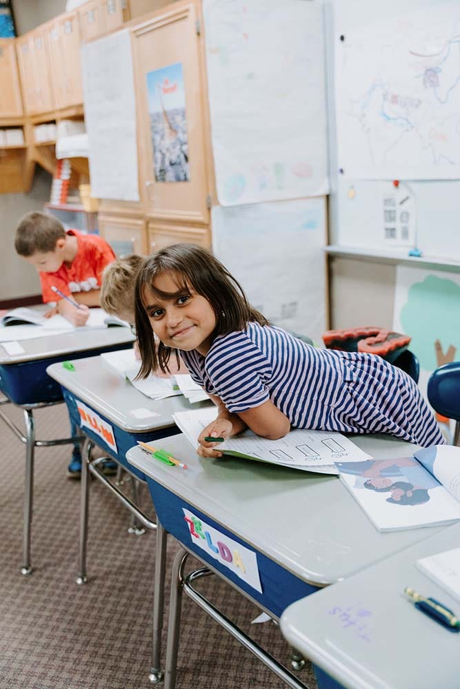 Students in classroom, sitting at tables, reading and coloring.