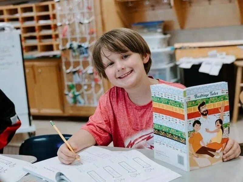 Students in classroom, sitting at tables, reading and coloring.