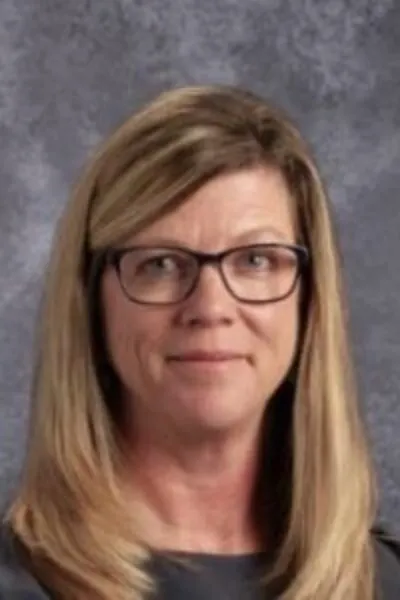 Smiling woman with straight blonde hair and glasses in front of a gray mottled studio background.