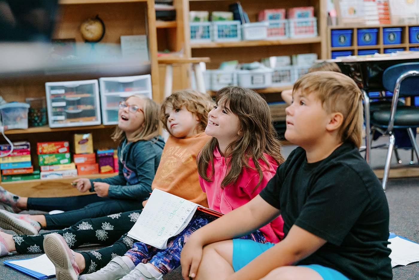 Students in classroom, sitting either on the floor or in a chair, participating in classroom activities