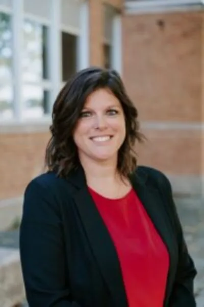 woman smiling in a black blazer and red blouse standing in front of a brick school building