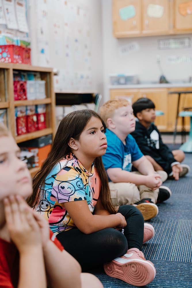 Students in a classroom engaging in classroom activities while sitting on the floor.
